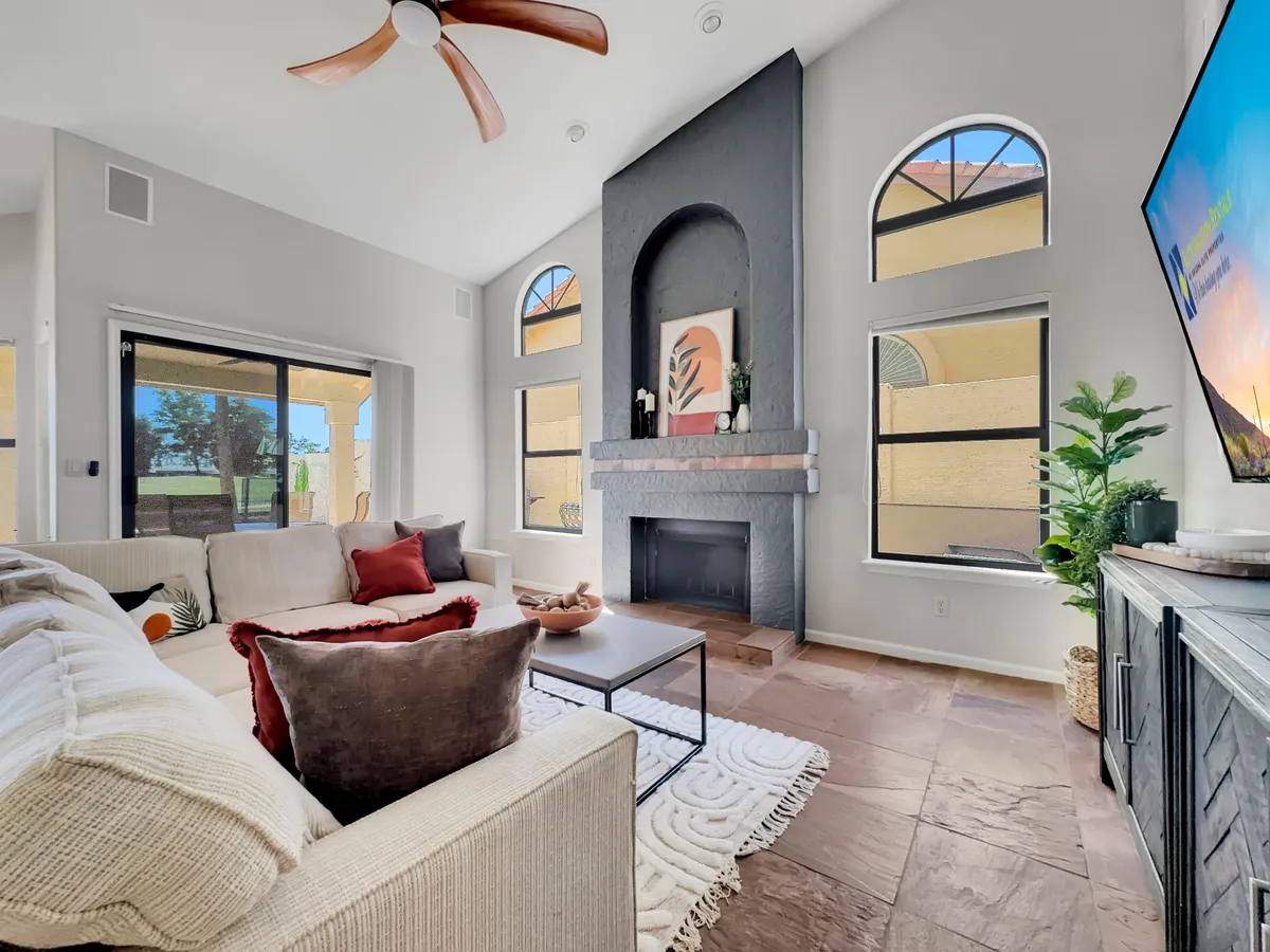A spacious, open-concept living room featuring a large sectional sofa facing a fireplace and a big-screen TV. Large windows frame beautiful lake views, and the space is accented by warm lighting and decorative plants. The plush seating is arranged to encourage conversation and togetherness.