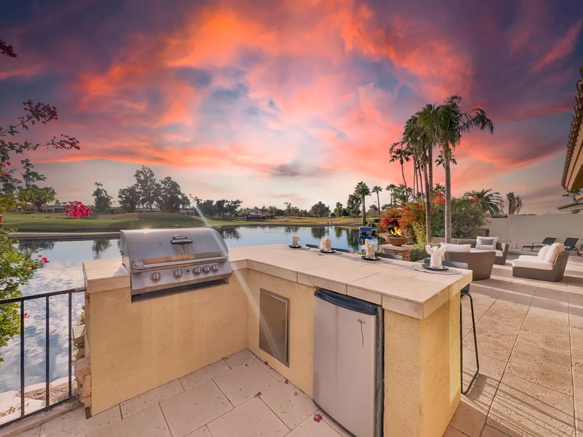 An outdoor kitchen area with built-in grill, set beside a tranquil lake. Palm trees and dramatic sunset colors create a stunning backdrop for entertaining and alfresco dining.