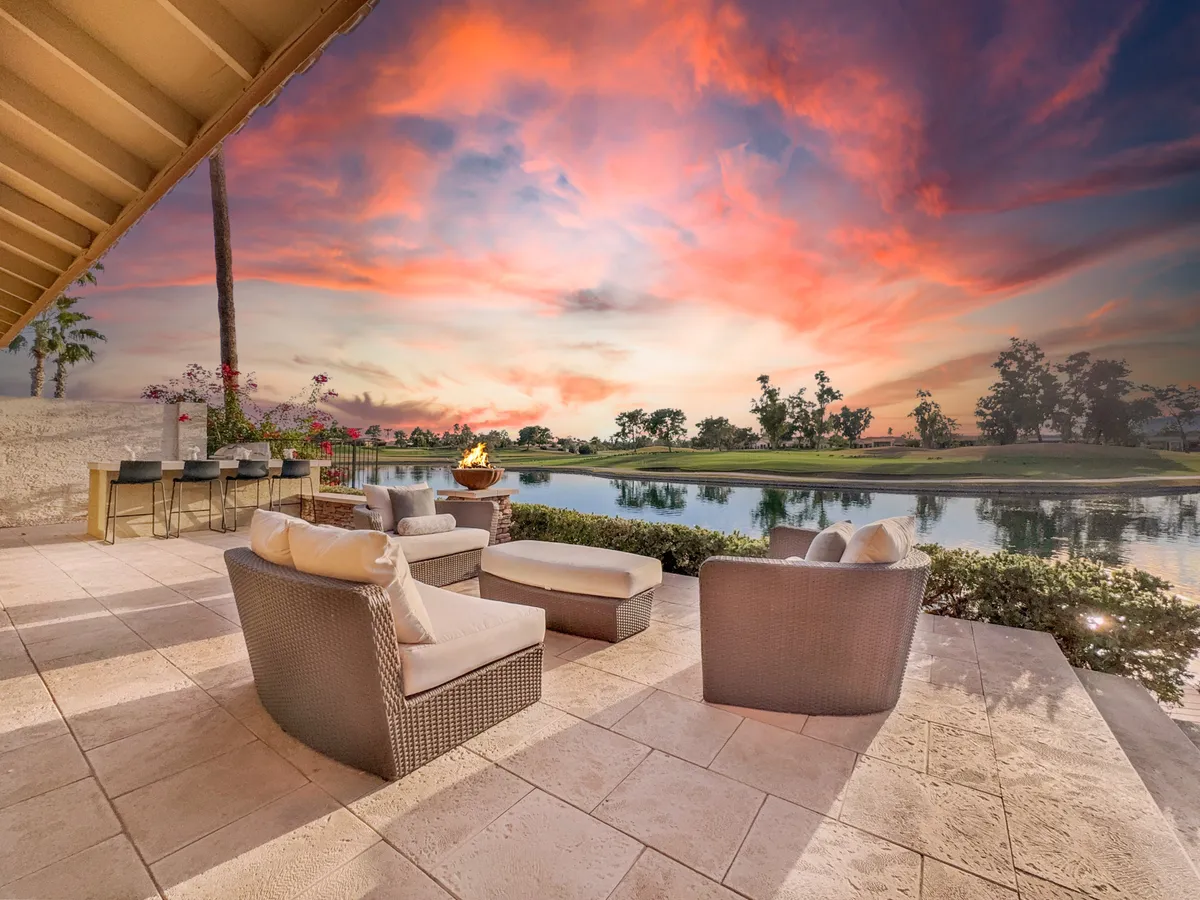 Cozy lounge seating surrounds a glowing fire pit, with an expansive water view and a dramatic sunset sky in the background. The setup suggests intimate gatherings, warmth, and relaxation at the water’s edge.
