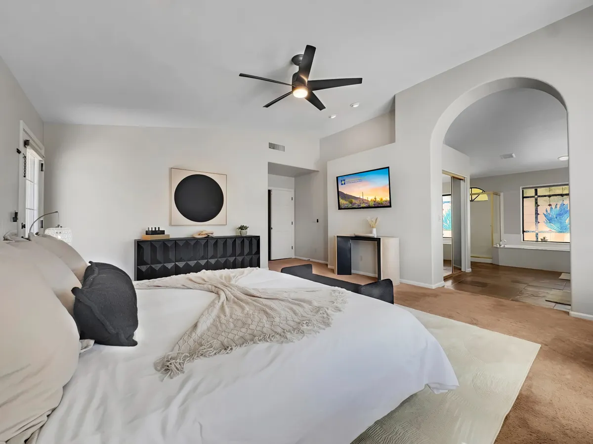 A king-size bed with plush, premium bedding is placed against a stylish black headboard, flanked by nightstands and a modern ceiling fan overhead. A large window lets in natural light, and a mounted TV is visible, creating a serene and inviting bedroom retreat.