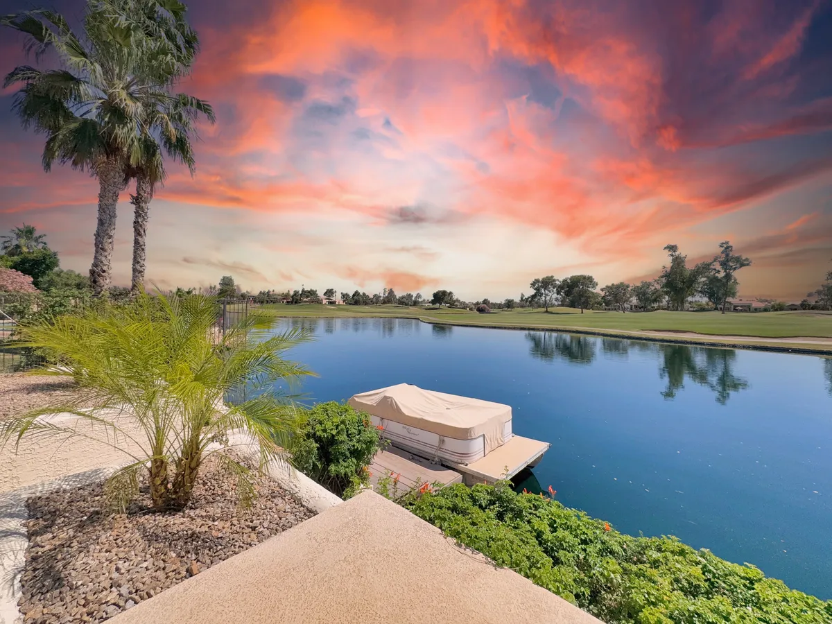 A scenic lakeside view at sunset, with palm trees reflected in calm water and a boathouse visible in the distance. The sky glows with warm colors, inviting thoughts of boating, fishing, and peaceful evening strolls by the water.