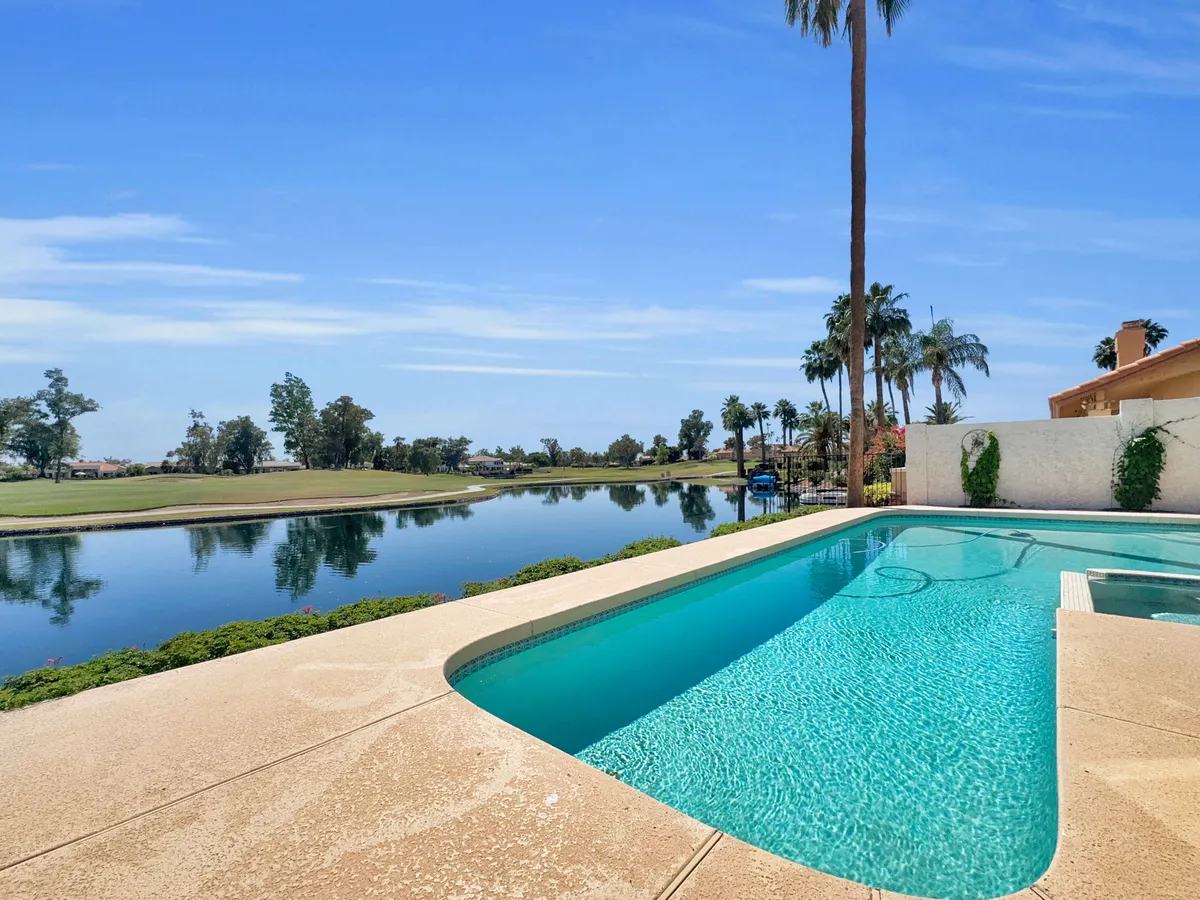 A tranquil backyard scene with a sparkling swimming pool, framed by palm trees and lush landscaping. Beyond the pool, the manicured green of a golf course is visible, creating a seamless connection between the home and the Ocotillo Golf Club. Sunlight reflects off the pool’s surface, evoking a relaxing, resort-like atmosphere.