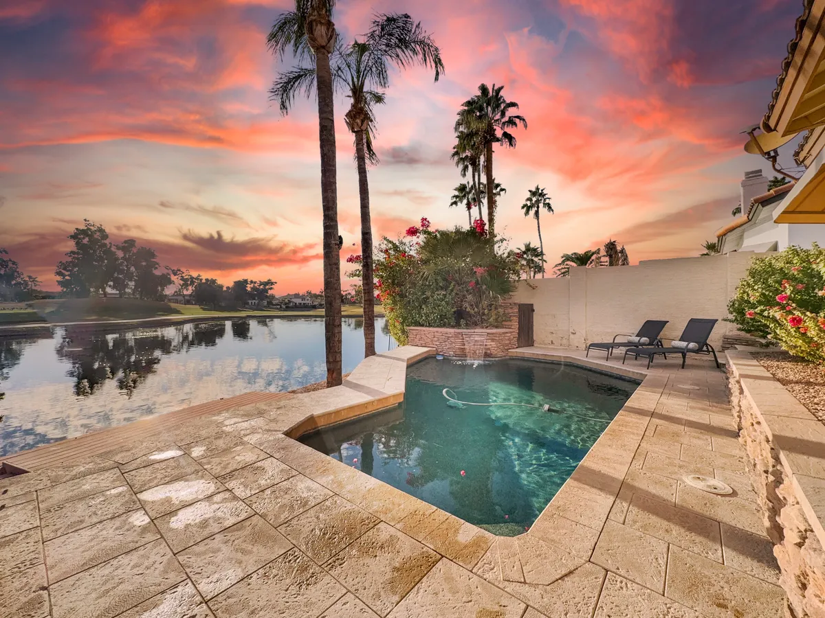 A serene outdoor pool area featuring sparkling blue water, tall palm trees, a spacious deck, and a glowing sunset sky reflected on the pool's surface. The pool is surrounded by lounge chairs and ample space for gatherings or relaxation.