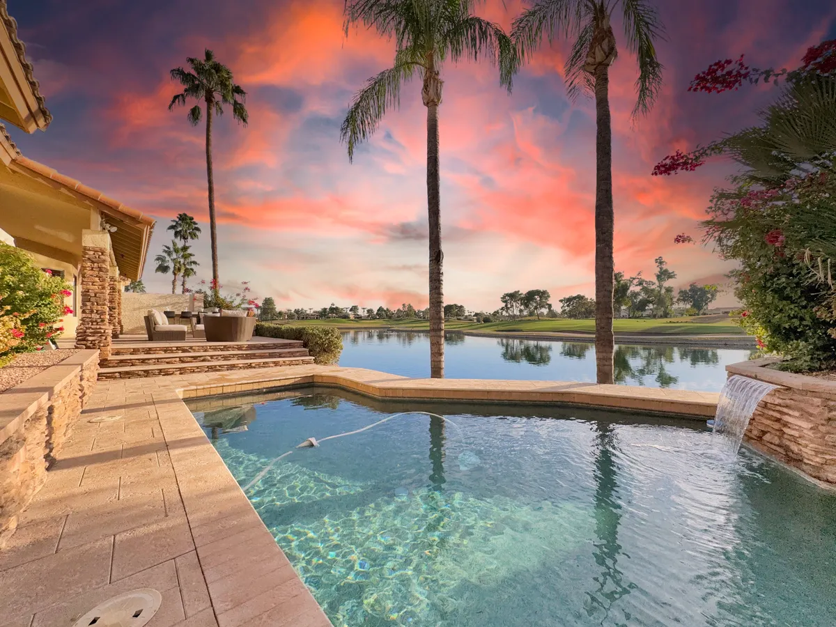 A luxurious outdoor space featuring a swimming pool with a waterfall, shaded seating areas, and a serene reflective lake beyond. Palm trees and lush greenery frame the scene, while the sun sets in the background, casting a warm glow over the entire area.